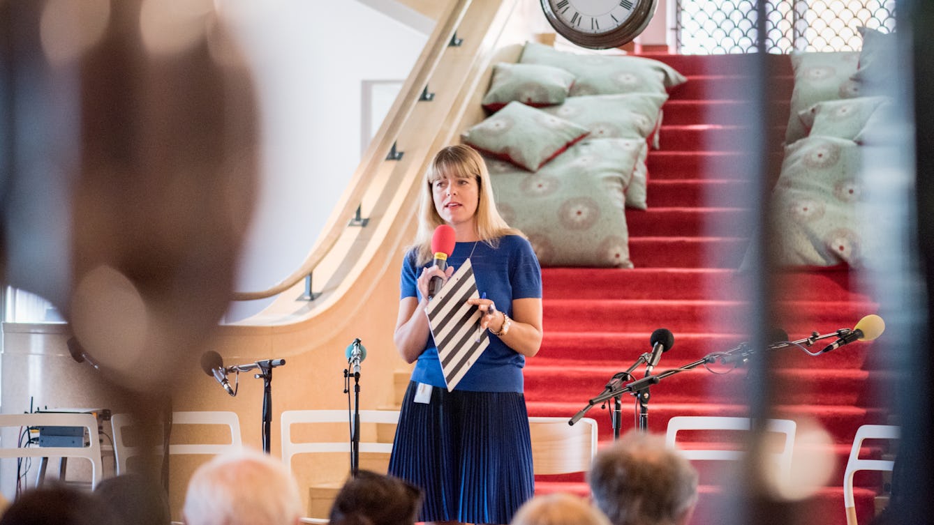Claudia Hammond standing in front of an audience in the Reading Room at Wellcome Collection. She is holding a microphone, behind her is a red carpeted staircase with cushions on it.