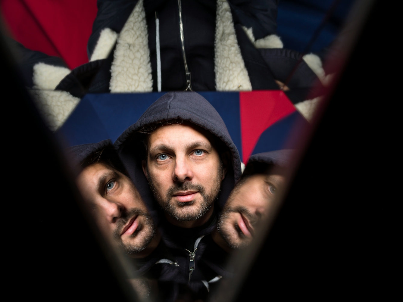 Photograph through a kaleidescope of the head of a man with his hood up.  The background is made up of blue and red fabric.