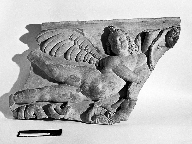 Black and white photograph showing a putto flying above a snake and a person whose face has been damaged.