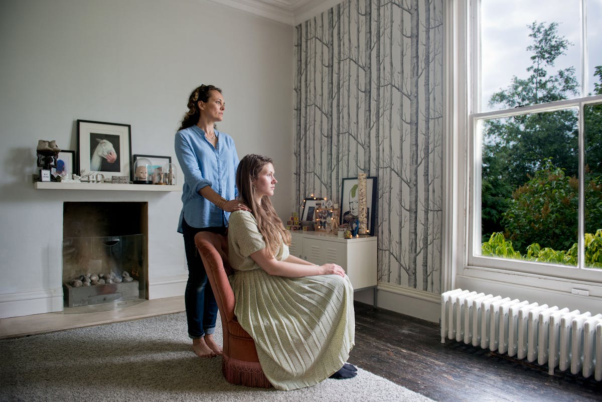 Photograph of a young woman and a mother figure in a domestic living room. The young woman is seated in a long dress and and the mother figure stands behind her, hands on her shoulders. They are both gazing out of the window.