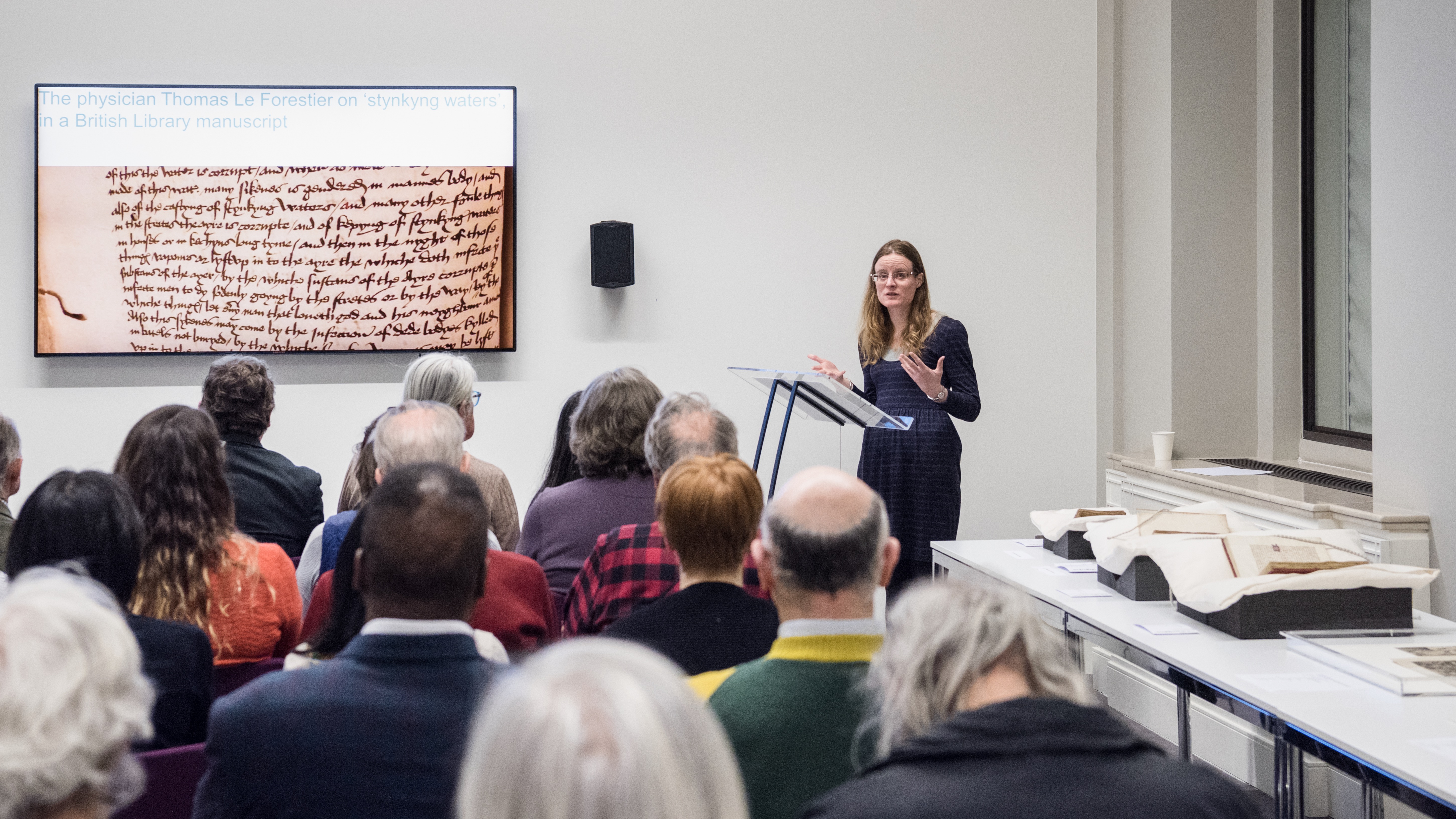 Photograph of a woman standing at a lecturn giving a talk to an audience. Beside her on the wall is a large screen showing her presentation slides. 