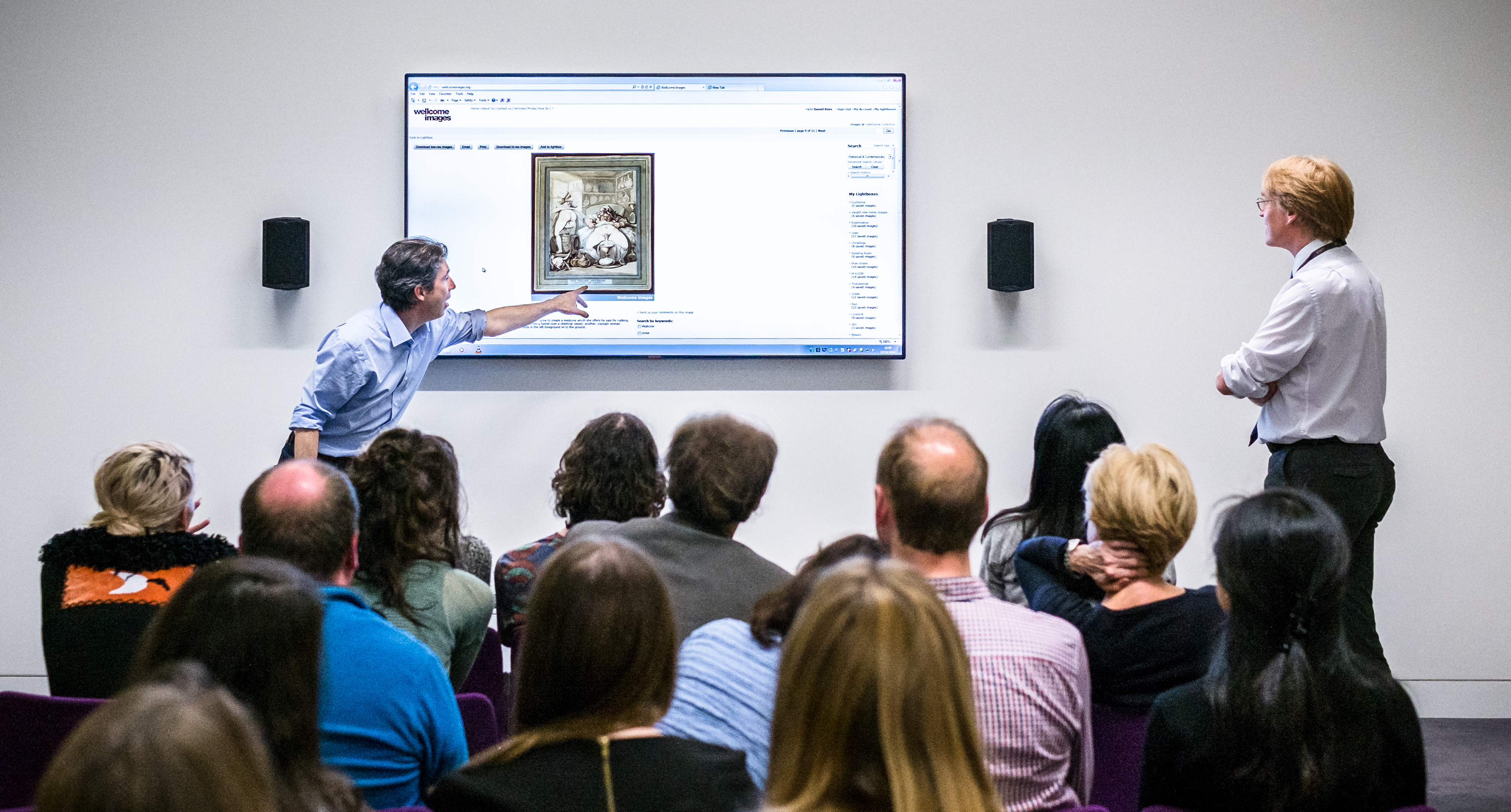 Photograph of a man giving a talk to an audience. He is pointing to a large screen besides him.