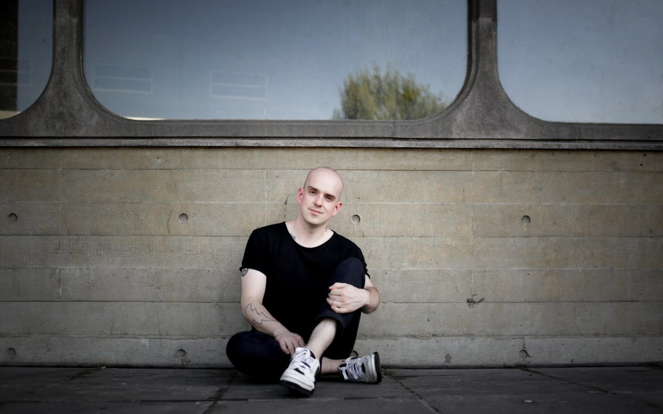 Photograph of a man dressed in black trousers and t-shirt, sitting on the pavement against a concrete wall containing large glass windows. 