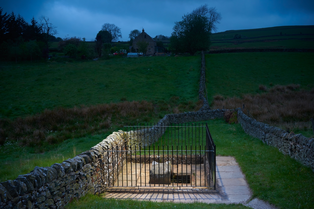 Photograph at dusk of a fenced off well, surrounded by dry stone walls and fields. IN the distance is a remote farm house on a hill. The well is spotlit.