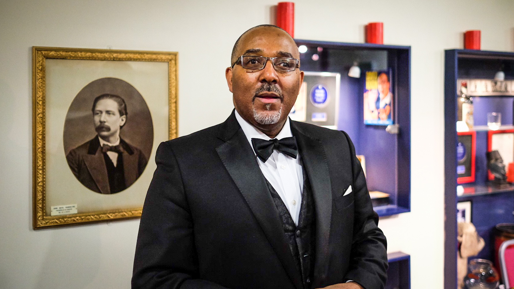 Photograph of a man in a dinner jacket and bow tie standing in front of a framed portrait hanging on a wall and several display cases.