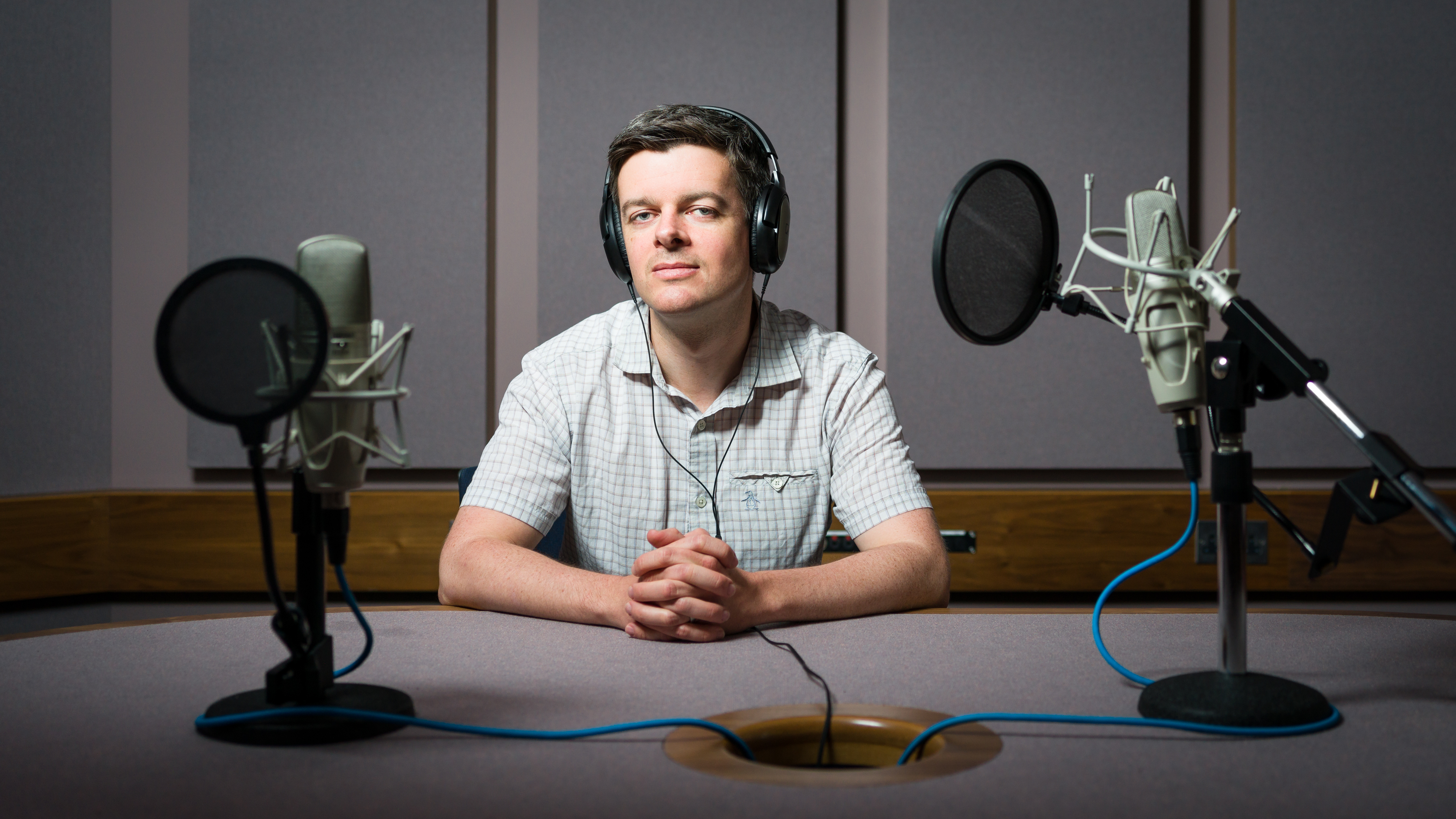 Photographic portrait of Dr. Adam Tierney sitting in a recording studio wearing headphones and surrounded by two microphones.