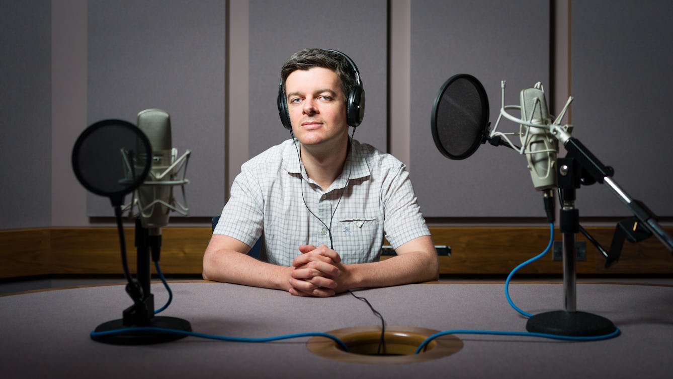 Photographic portrait of Dr. Adam Tierney sitting in a recording studio wearing headphones and surrounded by two microphones.
