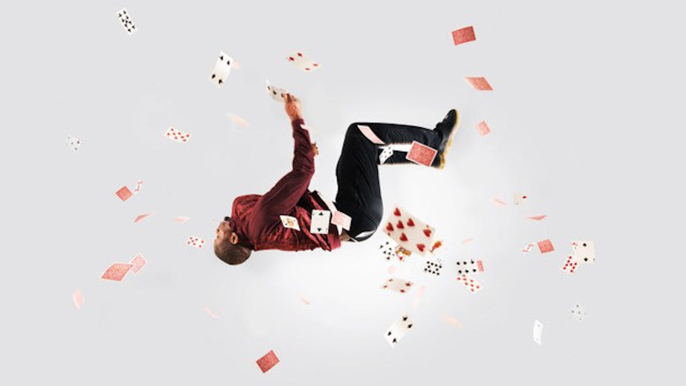 Photograph of a man backwards somersaulting through the air, wearing a red jacket and black trousers. He is surrounded by playing cards flying through the air around him.