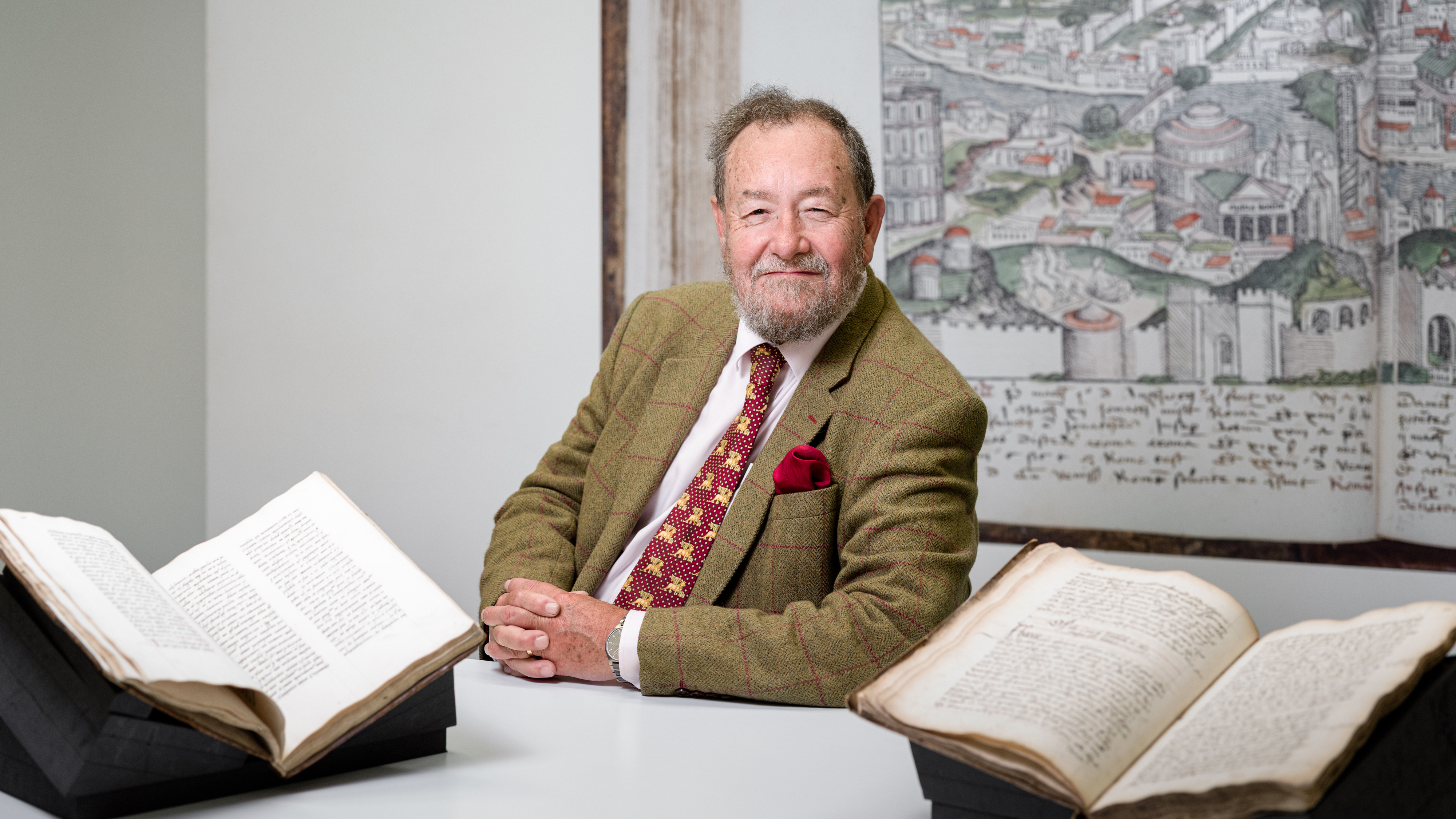 Photographic portrait of Dr Robert Hutchinson in the library at Wellcome Collection. In front of him sits two open manuscripts MS 7117 an English medical compendium c.1575 and MS 375 two surgical treatises of 1506. 