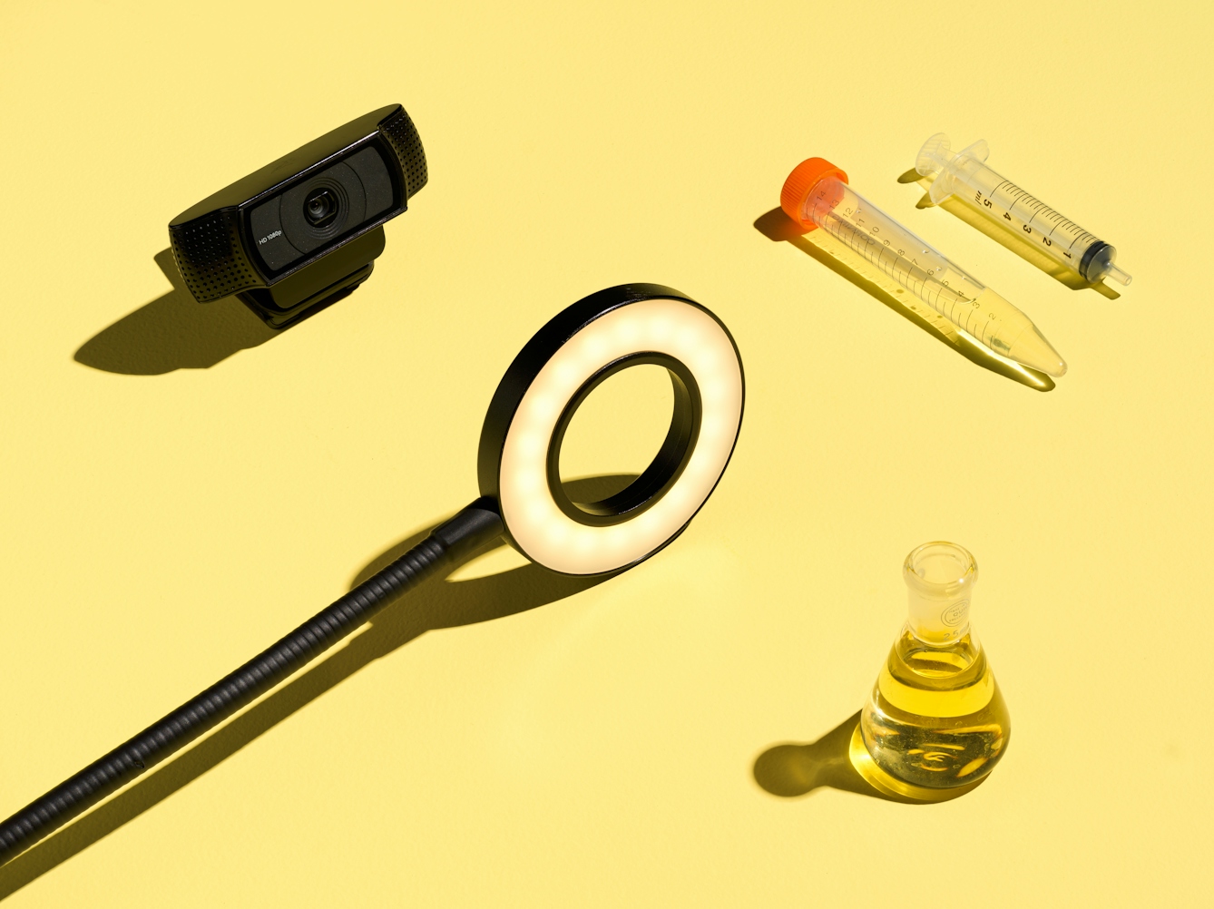 Photograph of 1 specimen bottle containing yellow liquid, 1 plastic specimen tube, 1 plastic syringe, a web cam ring light on a flexible arm and a web,. The whole scene has been photographed against a bright solid yellow background.