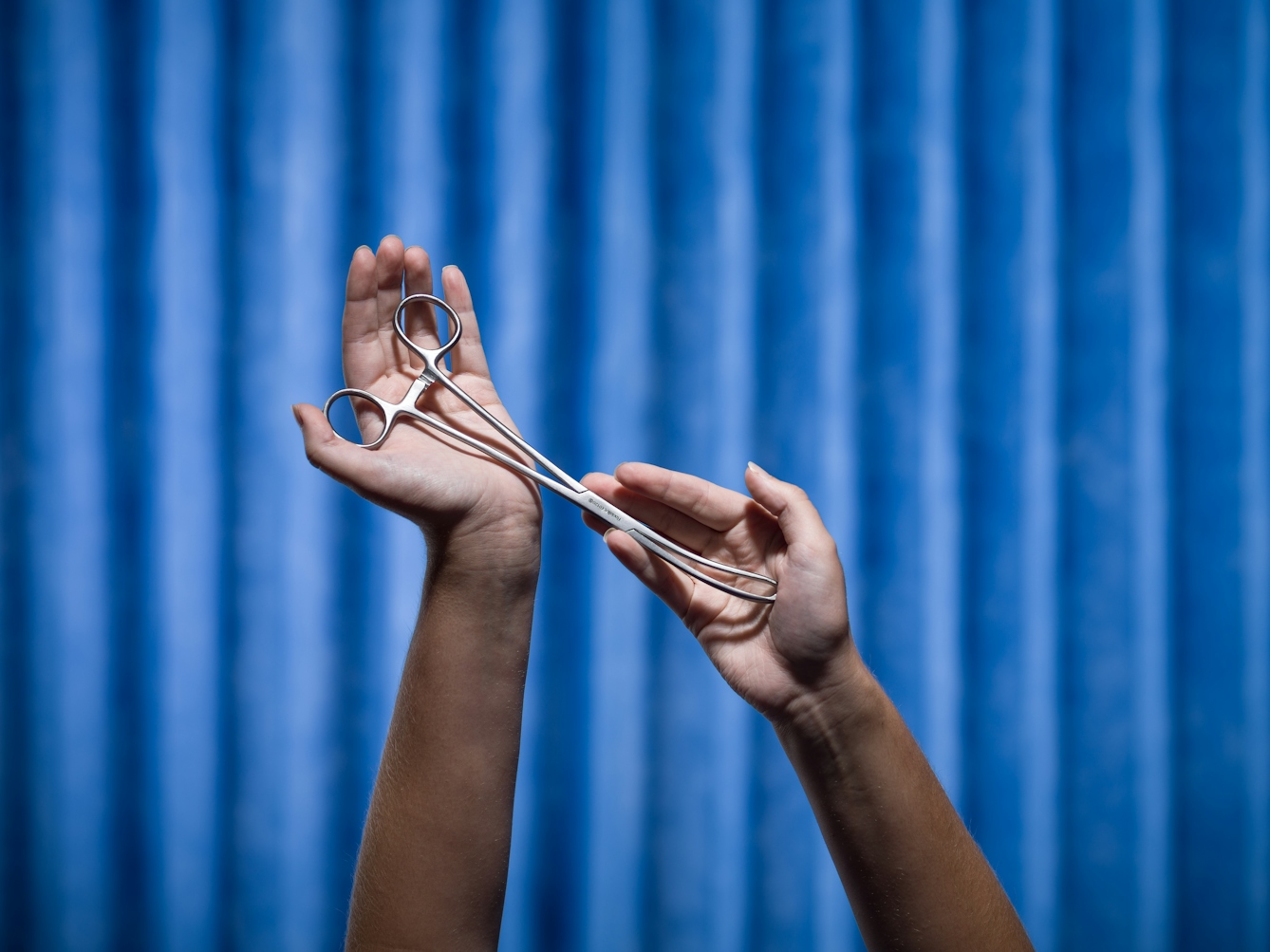 Photograph of a pair of hands cradling a set of forceps. Photographed against a background of blue antibacterial clinical curtains.