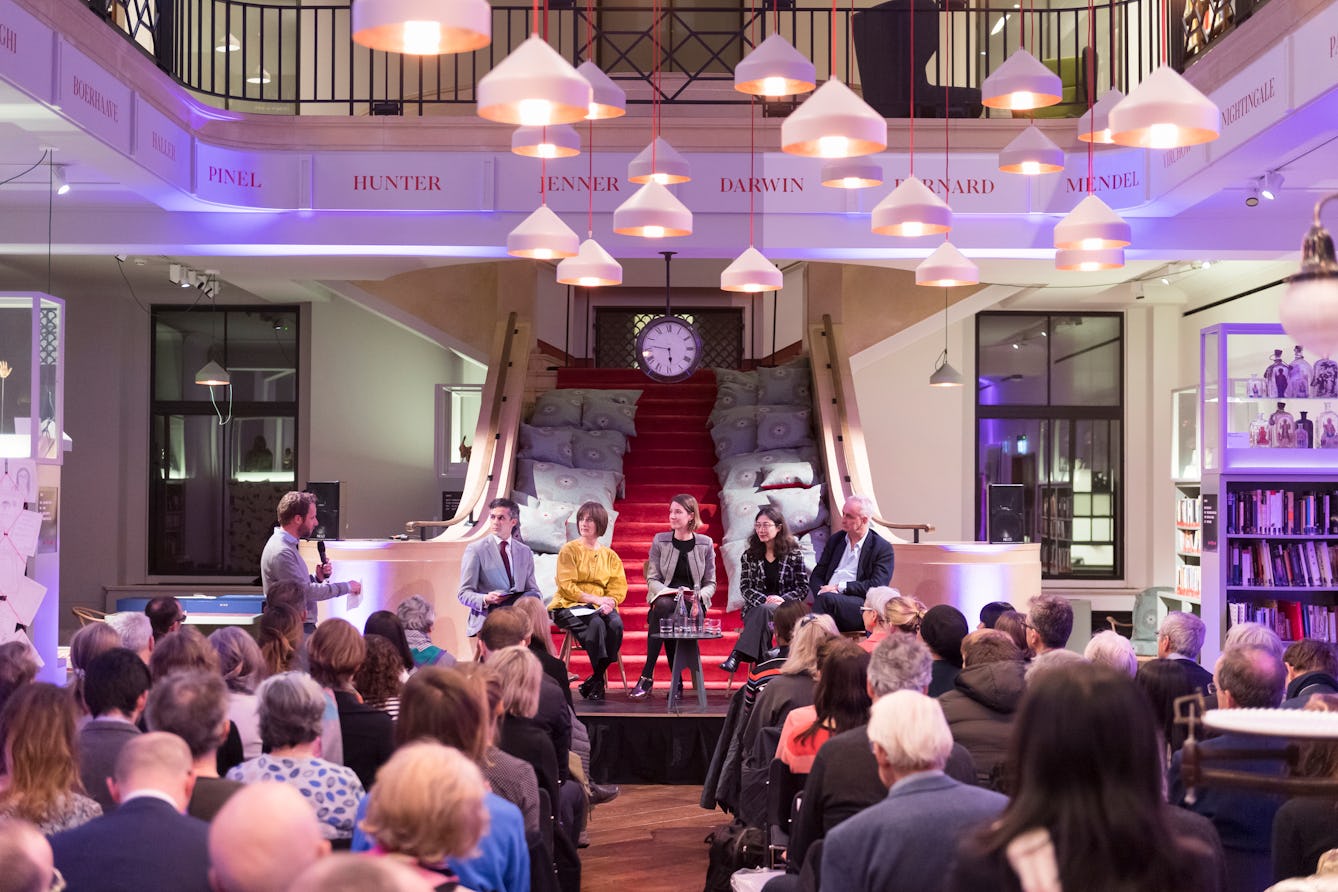 Photograph of five panellists sat on a raised stage in front of an audience. Behind them is a red carpeted staircase. The walls are bathed in a purple wash of up-lighters.