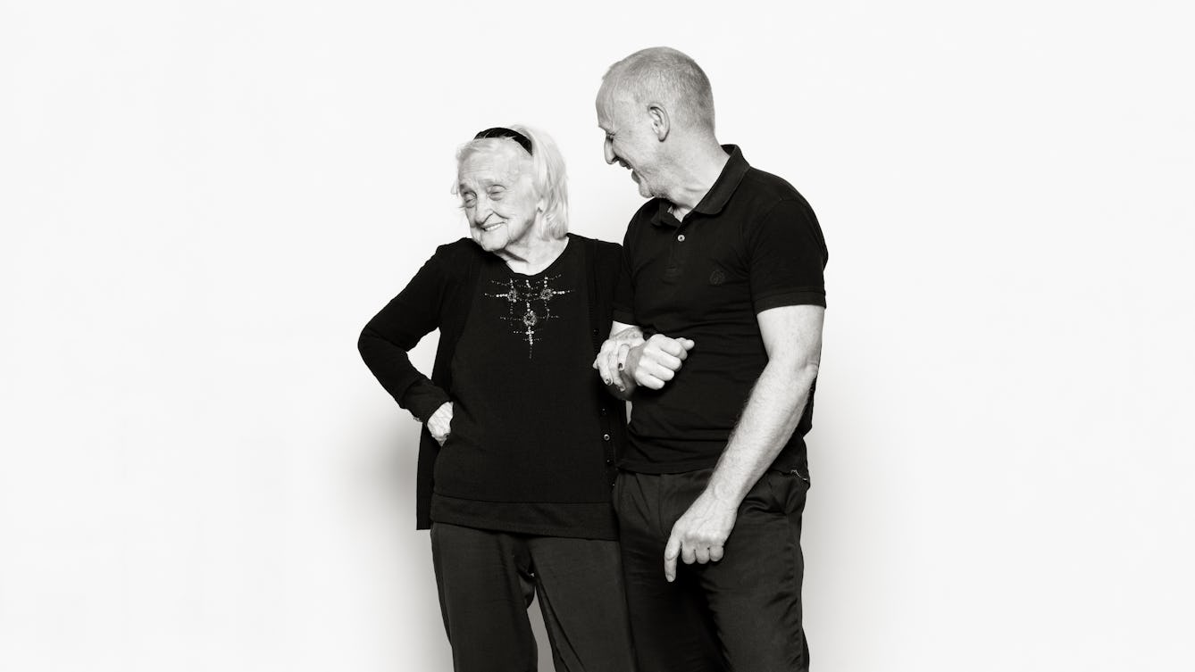 Photographic portrait of an elderly lady linking arms with a male carer against a white background.