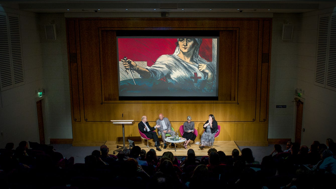Photograph of a dark auditorium showing the backs of the audience's heads and four panel members seated on a small stage, in-front of a large screen. On the screen is a still from a film.