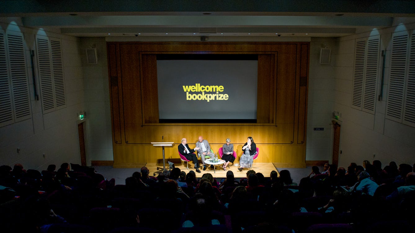 Photograph 4 people seated on the stage as part of a panel discussion in the Henry Wellcome Auditorium, with a projector screen behind them and an audience in front.