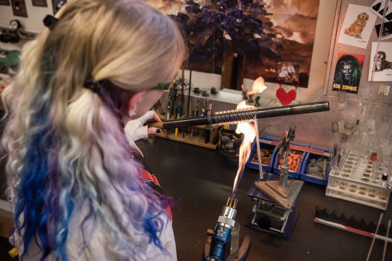 Photograph from behind a woman with blonde hair streaked with blue and purple, holding a metal rod wrapped with a glass spiral over the flame of a blowtorch.