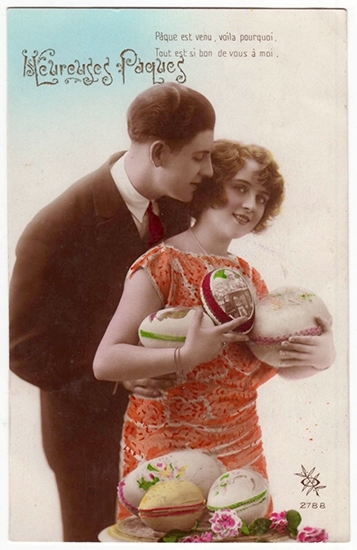 A young woman in an orage dress stands infront of an table with three large Easter eggs and scattered flowers on it, while she holds three large decorated easter eggs in her arms and a young man in a smart suit embraces her from behind,