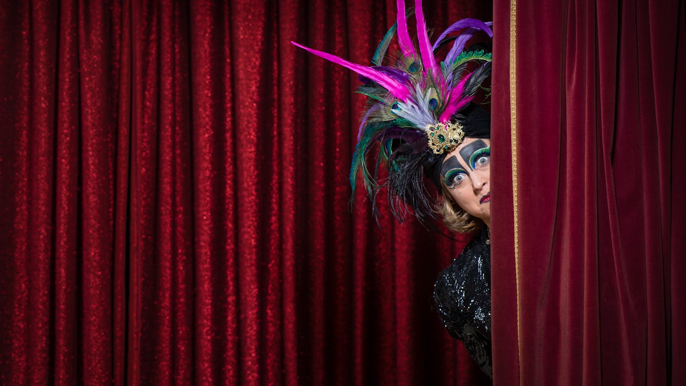 Photograph of a woman wearing a colourful feathered headdress and face makeup, peeping around the red velvet curtain of a theatrical stage set.