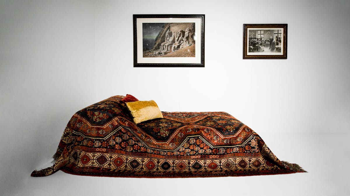 Photograph showing a Persian rug draped over a couch in a white exhibition setting. On the wall behind the couch are 2 framed pictures.