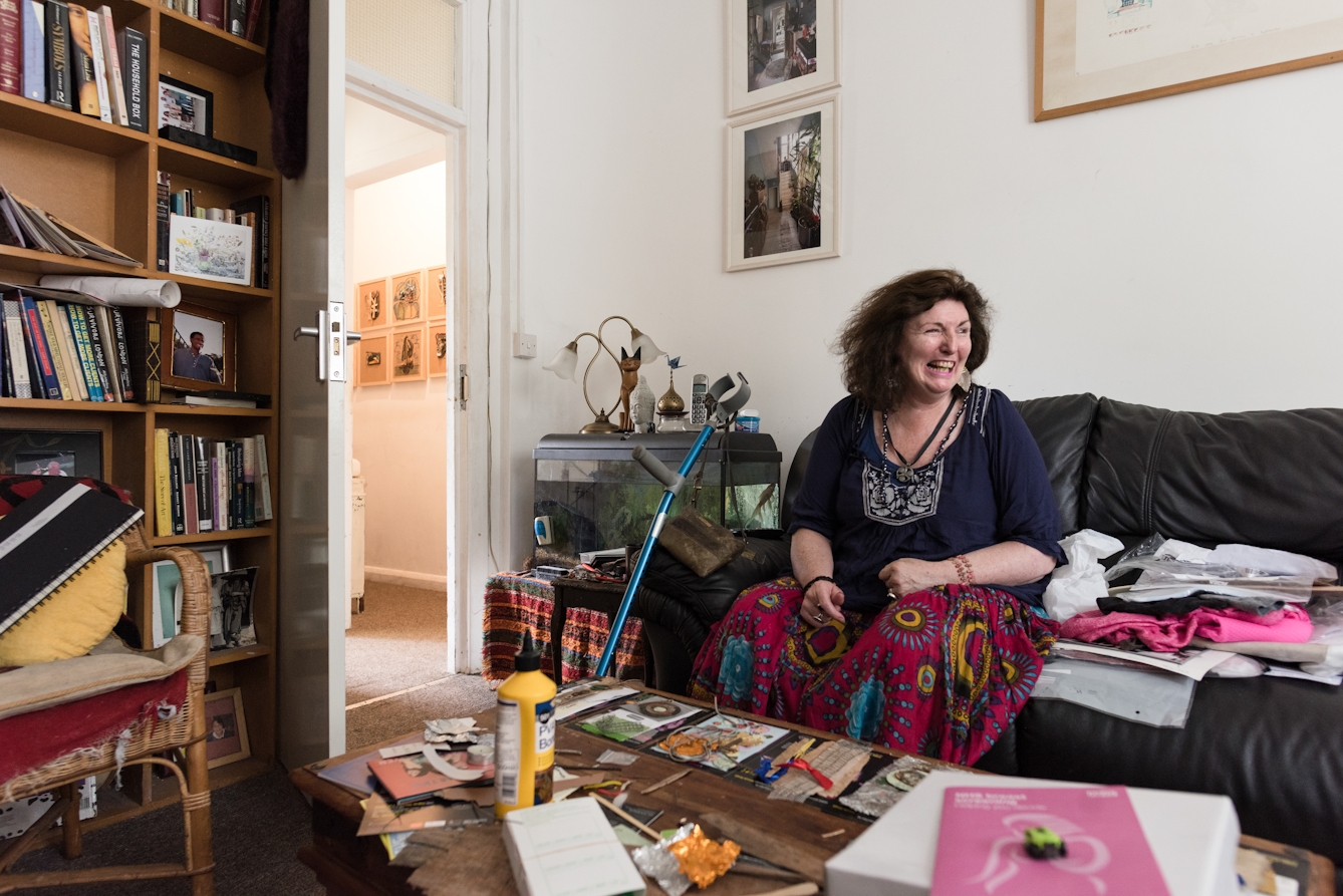 Photograph of a woman sitting on a sofa in her home laughing. She is surrounded by her artworks and a crutch.