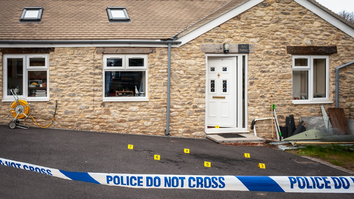 Photograph of the front of a house with the front door slightly open and forensic crime scene numbered yellow markers on the ground. In the foreground is a police cordon with 'Police Do Not Cross' written across the tape.