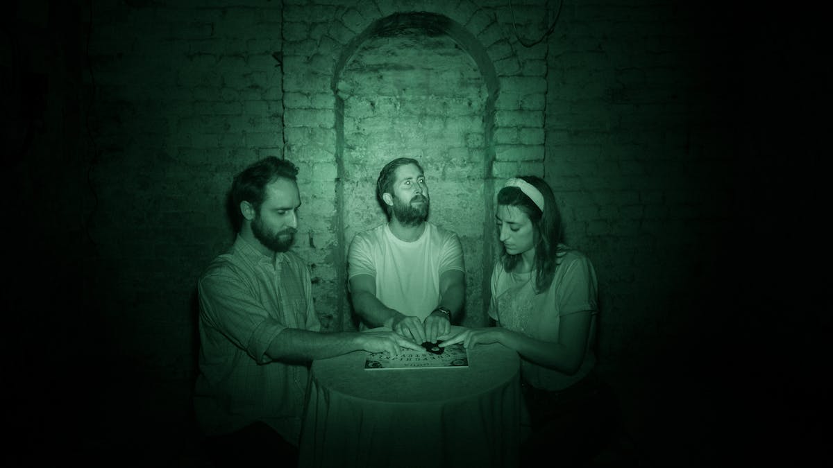 Photograph of three people sat at a round table in a cellar. Behind them is a white brick wall with an arch. Their hands rest on the planchette of a ouija board. The image is toned green as a result of being made under infrared light.