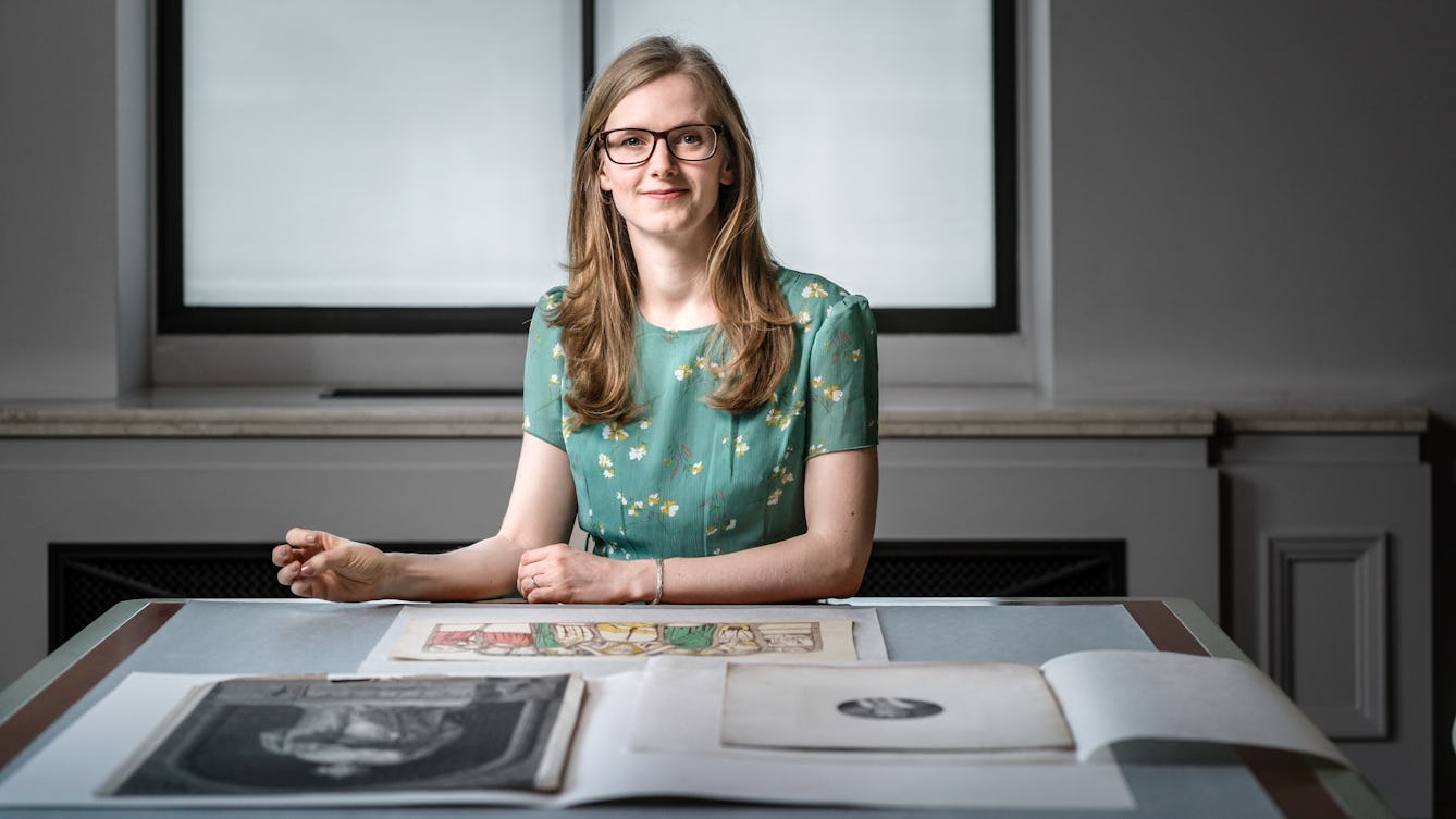 Photograph of Lauren Johnson sitting behind a desk in the Reading Room at Wellcome Collection, with a selection of collection items in front of her.