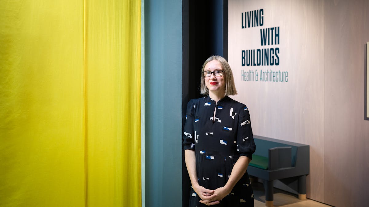 Photographic portrait of Emily Sargent, curator of the exhibition Living with Buildings at Wellcome Collection. Emily is leaning against a pillar within the exhibition space, with a hanging yellow drape to the left and the title of the exhibition to the right.