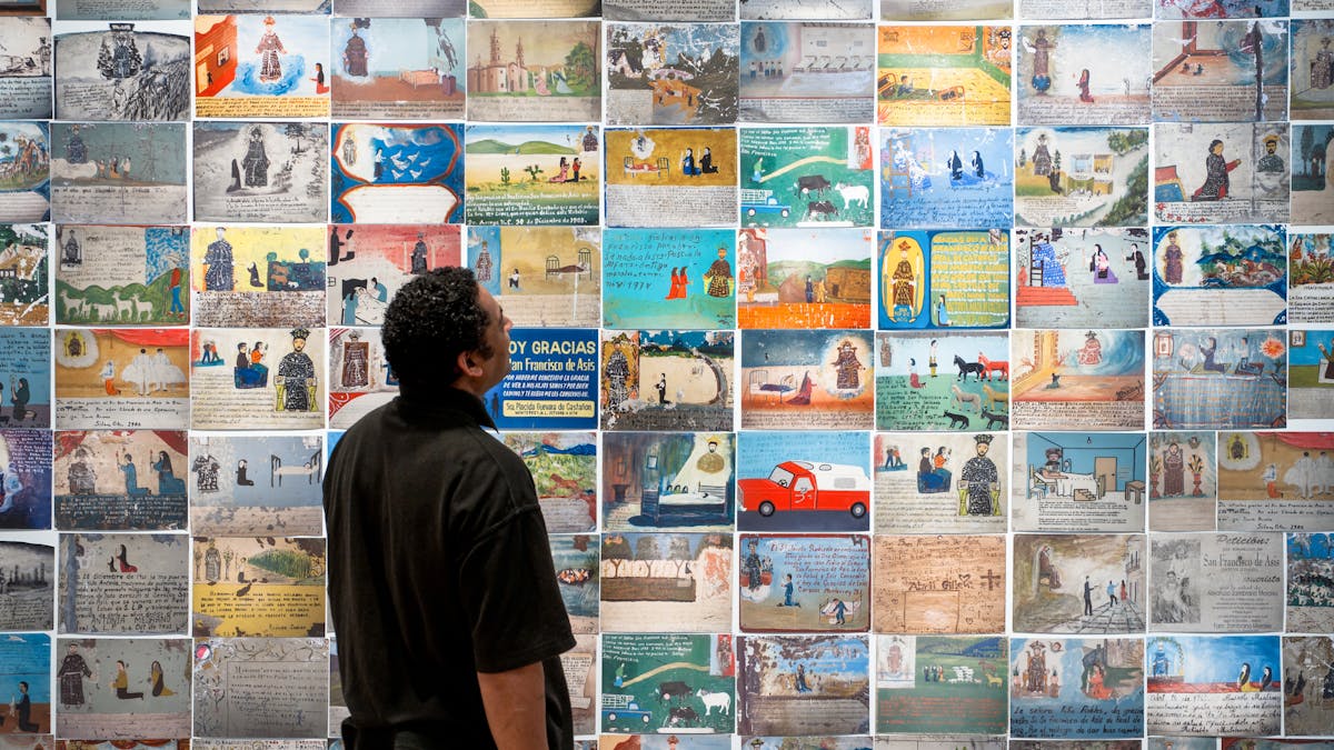 Photograph of a man looking at a wall of colourful Mexican votives, part of the exhibition Infinitas Gracias: Mexican miracle paintings at Wellcome Collection.