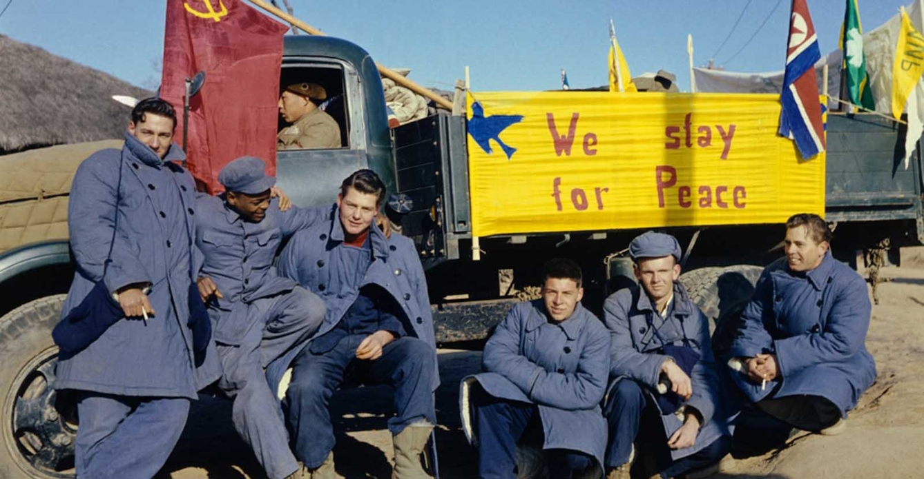 A group of black and white soldiers sit in front of a truck bearing a Chinese flag, and a banner declaring 'We Stay For Peace'.