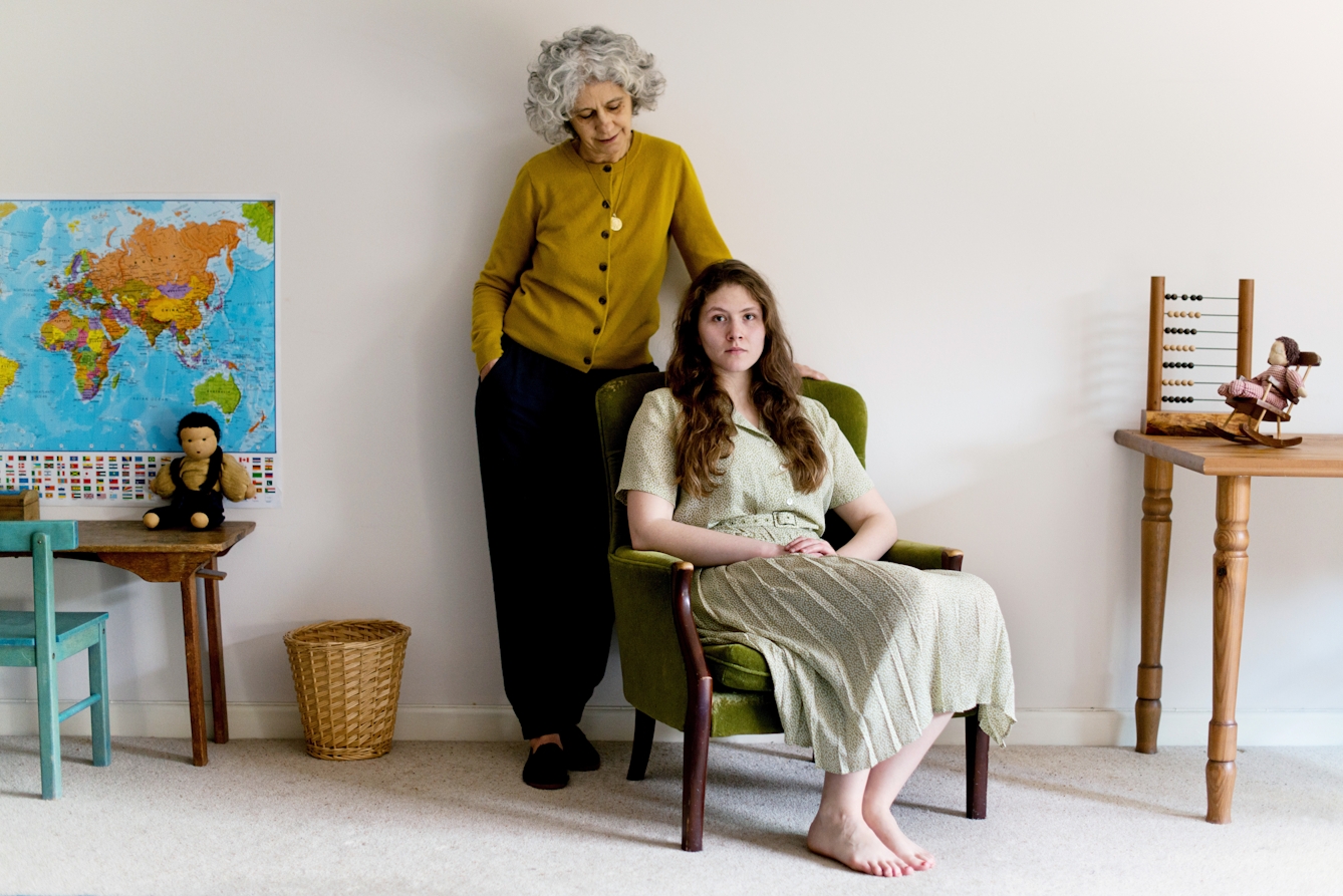 Photograph of a young woman and a mother figure in a child's bedroom. The young woman is seated in a long dress and and the mother figure stands behind her, hand on the back of her chair. The young woman is looking to camera, the mother figure is looking at her.
