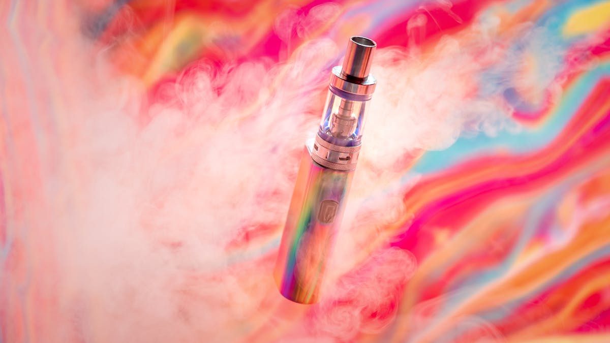 Photograph of an e-cigarette, against a colourful abstract background surrounded by wisps of smoke.