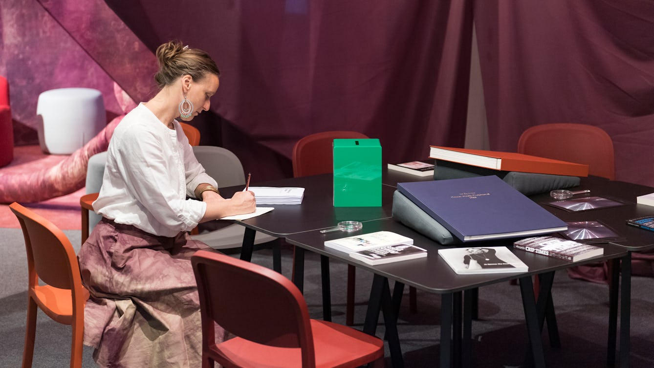 Photograph of a person sitting at a table within the Misbehaving Bodies exhibition at Wellcome Collection. They are holding a pencil and writing on a piece of paper. There are several books scattered around the table. 
