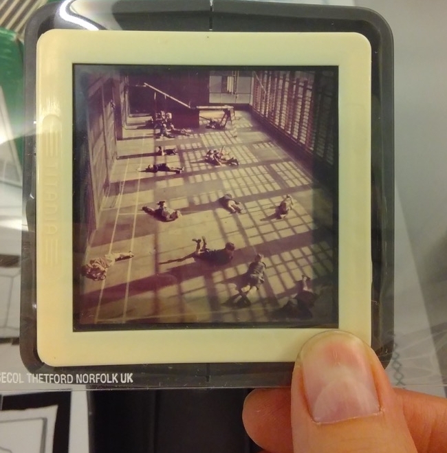 A close-up photo of someone holding a black and white slide between forefinger and thumb; the slide shows people lying on the floor of a large room or gymnasium.