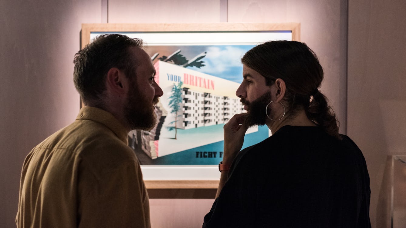 Photograph of two young men in conversation, standing in front of a poster exhibited as part of the Living with Buildings exhibition at Wellcome Collection.