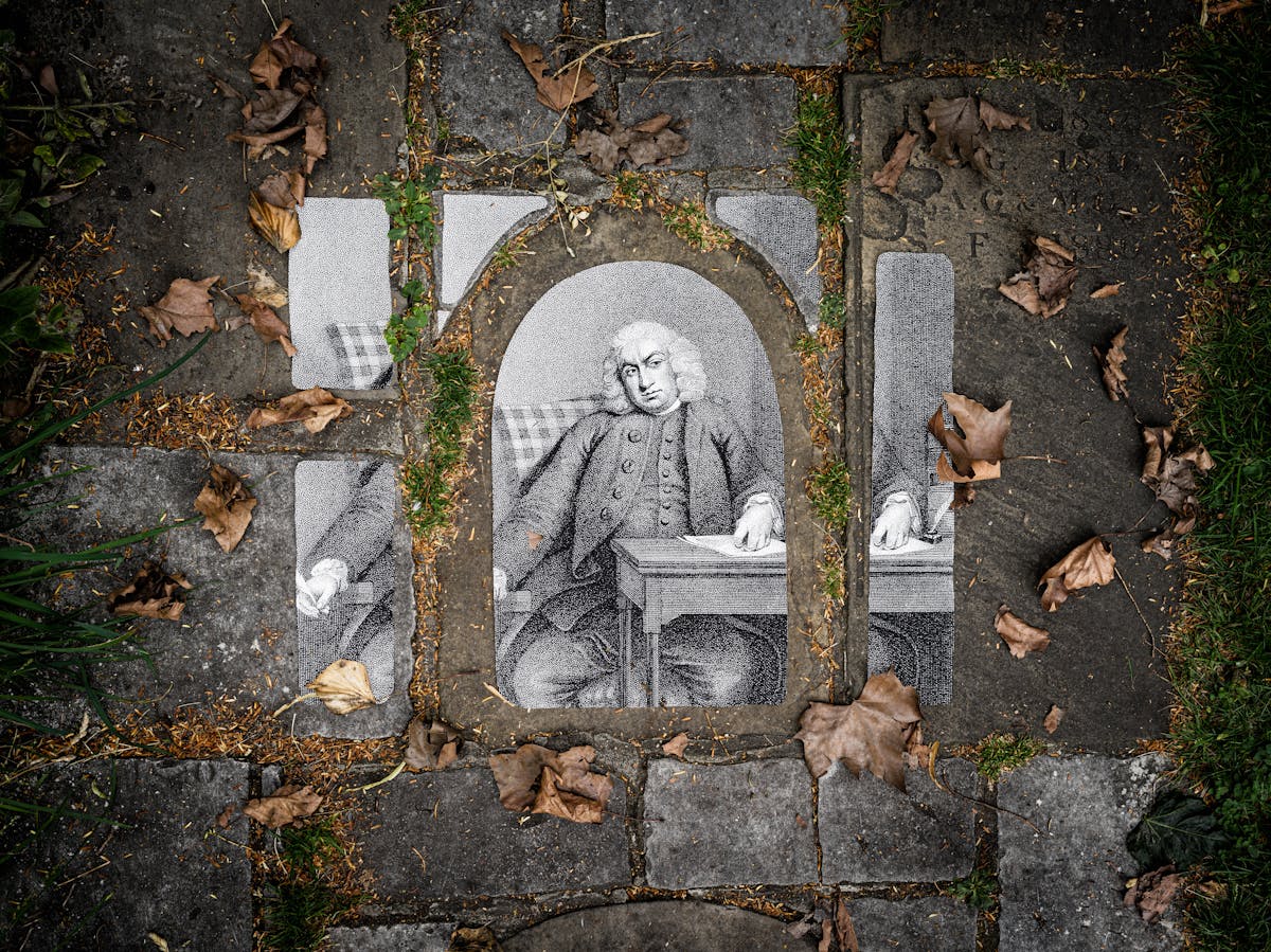 Photograph of an historical portrait drawing of Samuel Johnson placed on a path made out of paving slabs of different shapes and sizes. The portrait is cut into different shapes and sizes to match the paving stones beneath. Grass is growing between the slabs and leaves cover the ground.