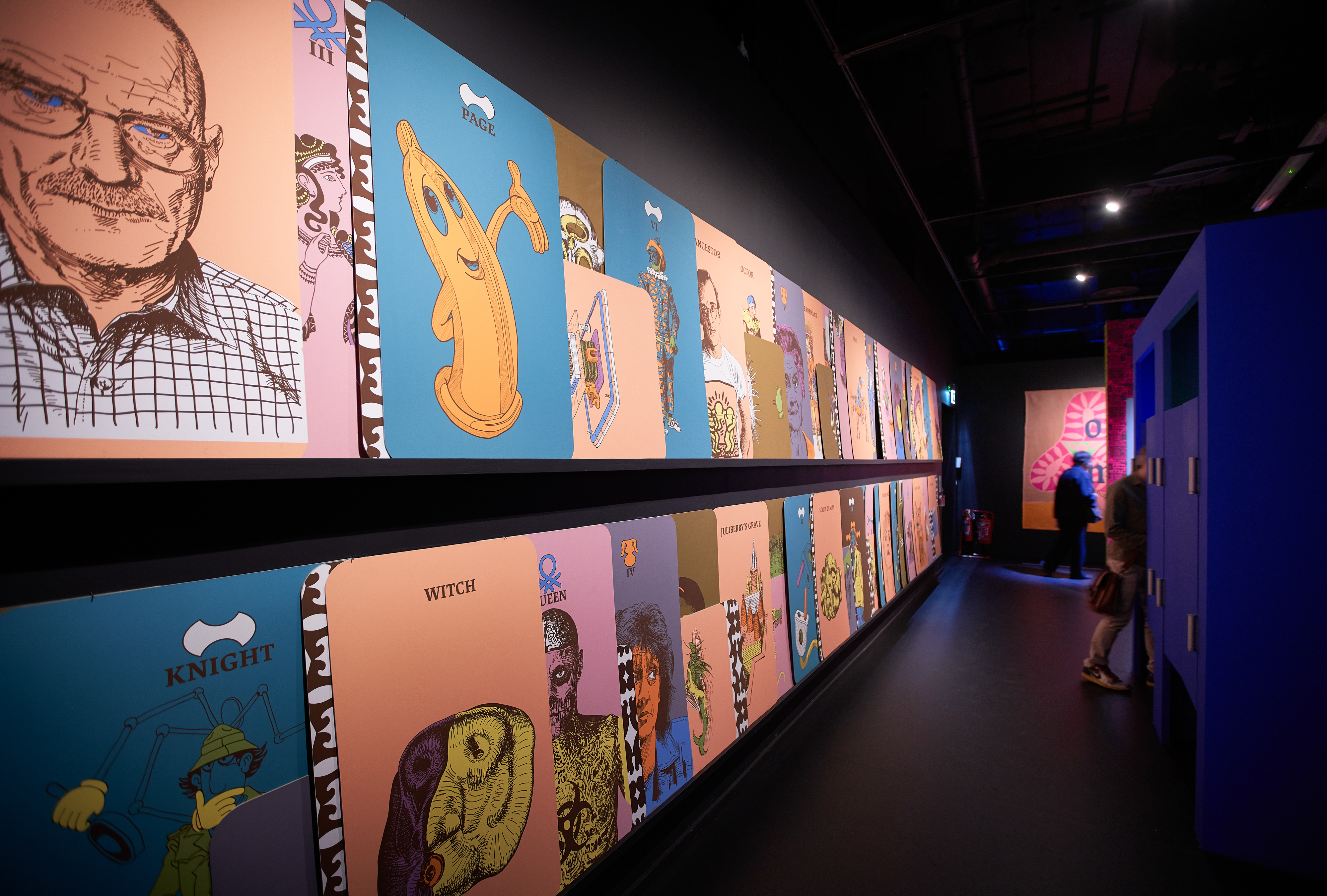 Photograph of a wall of colour large artworks, leaning against the wall on two long shelves which disappears into the distance. In the background are two gallery visitors.