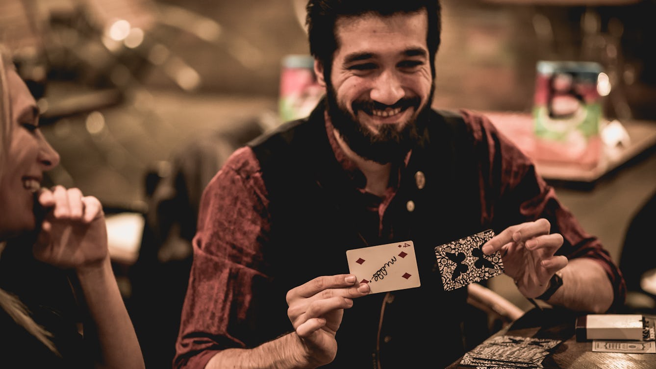 Photograph of a man with a beard holding up two paying cards as part of performing a trick to a small audience.