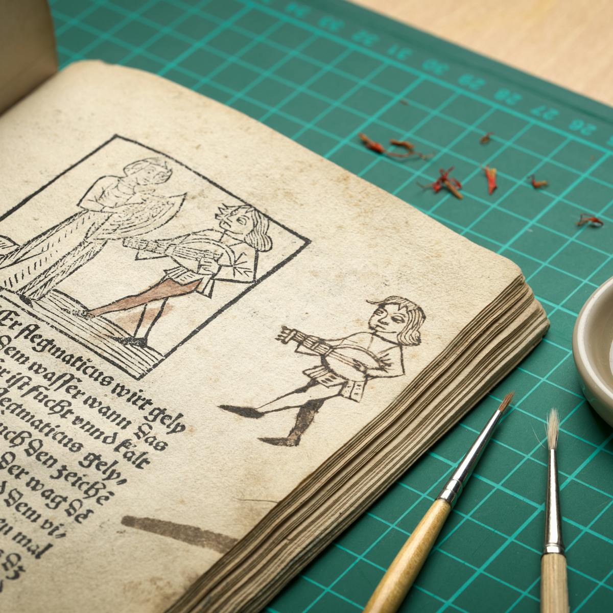 Photograph of an early printed book open as a page where a previous owner has had a go at copying an illustration of a man playing a guitar like instrument. Next to the open book are a couple of paint brushes and a small dish with saffron leaves on it.