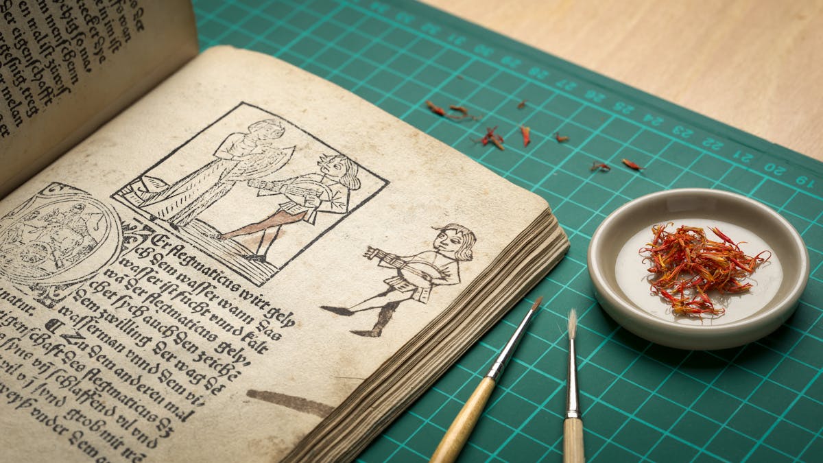 Photograph of an early printed book open as a page where a previous owner has had a go at copying an illustration of a man playing a guitar like instrument. Next to the open book are a couple of paint brushes and a small dish with saffron leaves on it.