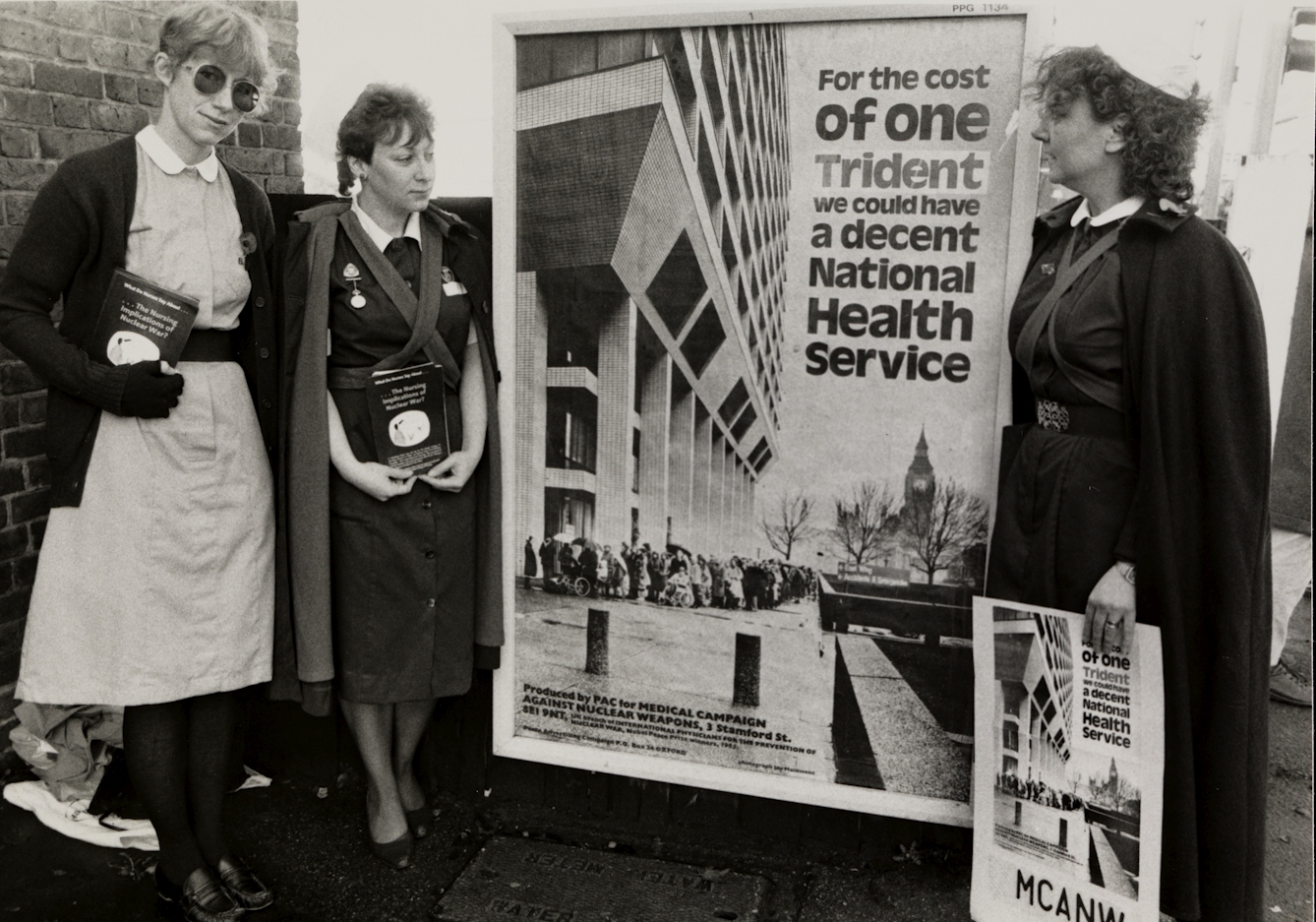 Black and white photograph of three nurses in uniform protesting about spending money on nuclear weapons rather than healthcare. They stand either side of a poster that reads "For the cost of one Trident we could have a decent National Health Service."