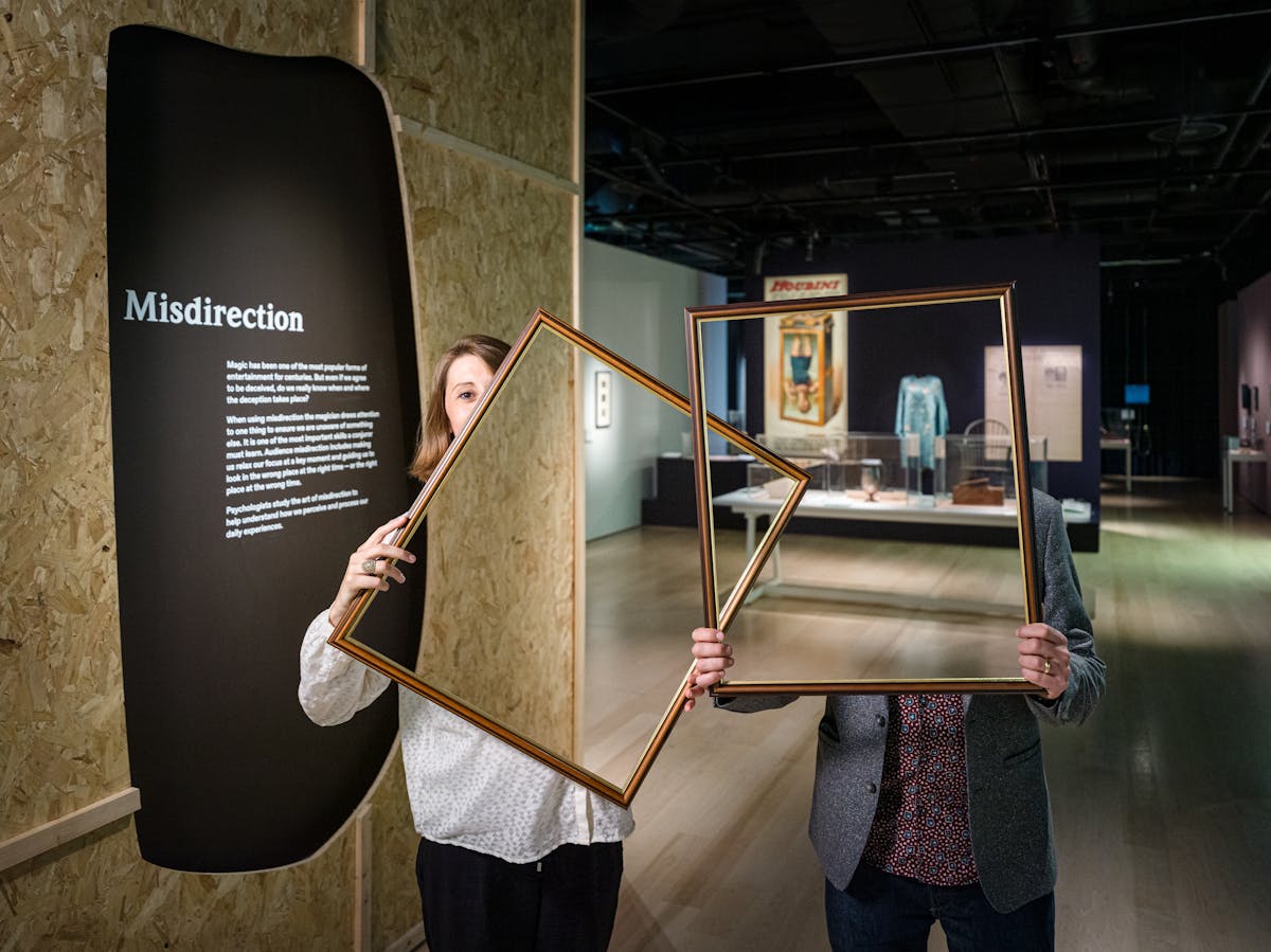 Photograph of a woman and a man in a dark gallery space holding up picture frames in front of their bodies and parts of their faces. Within the picture frame you can see straight through them, as if their bodies and faces have disappeared. In the background you can see a large historical poster and exhibitor display cases. On the left is a text panel titled, "Misdirection".