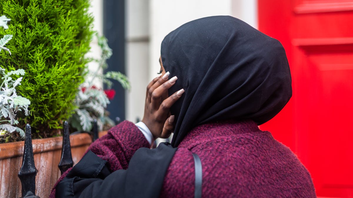 Photograph of a woman wearing a black head scarf facing away from the camera, with her hand held up against her cheek. In the background is the red front door of a house, some black railings and plants in a window box.