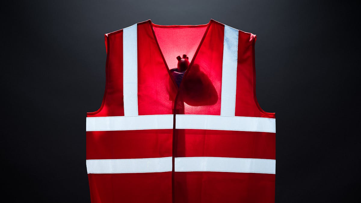 Photograph of a bright red high visibility vest against a dark grey background. The vest is lit from behind revealing the silhouette of a model of a human heart. The reflective strips on the front of the vest are bright with white light.