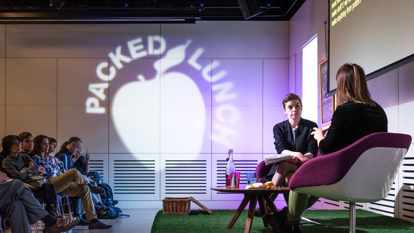 Photograph of two women having a conversation on a small stage. In the background are the audience. Projected onto the far wall is the silhouette of an apple with the words Packed Lunch circling the top of the apple. 