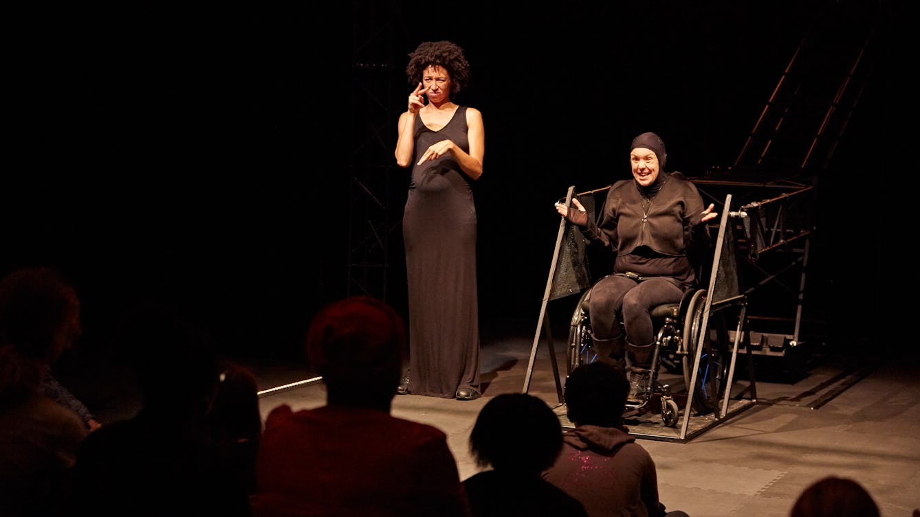 Jess Thom performing on a stage with a BSL interpreter, Charmaine Wombwell. The backdrop is black. Jess and Charmaine are both wearing black and are in front of an audience. 