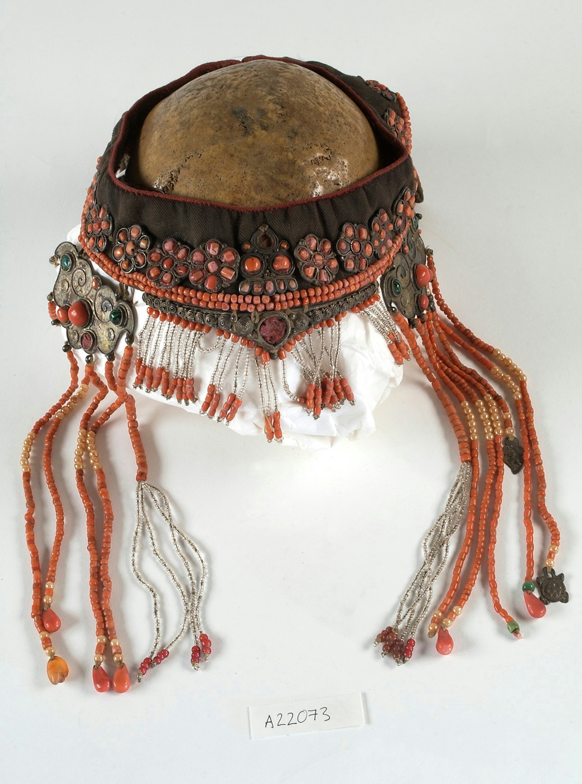 A photograph of a headdress made from brown material, decorated with clear, pink-red and green beads, and decorative metalwork. 