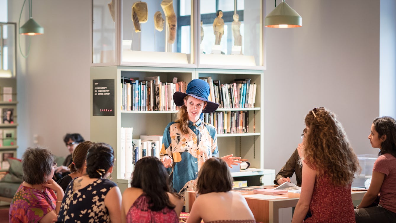 Photograph of a young woman wearing a large hat talking to a group of seated people. Behind her is the book shelves and exhibits displayed in the Reading Room at Wellcome Collection.