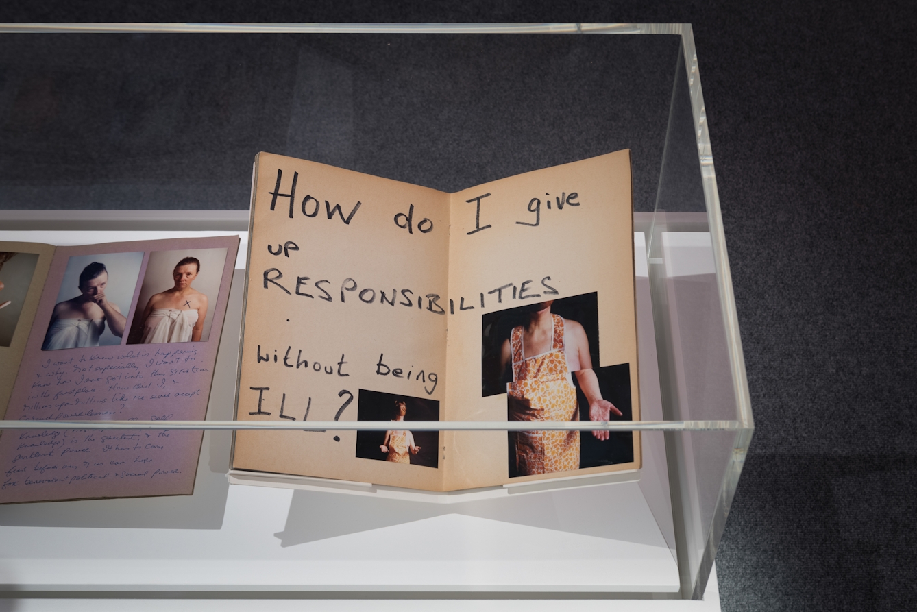 Photograph of a section of a gallery display case showing the open pages a scrapbook. The open page shows three photographs of a woman wearing a yellow pattered apron, with her hands outstretched. On the page is written the words, 'How do I give up responsibilities without being ill?'