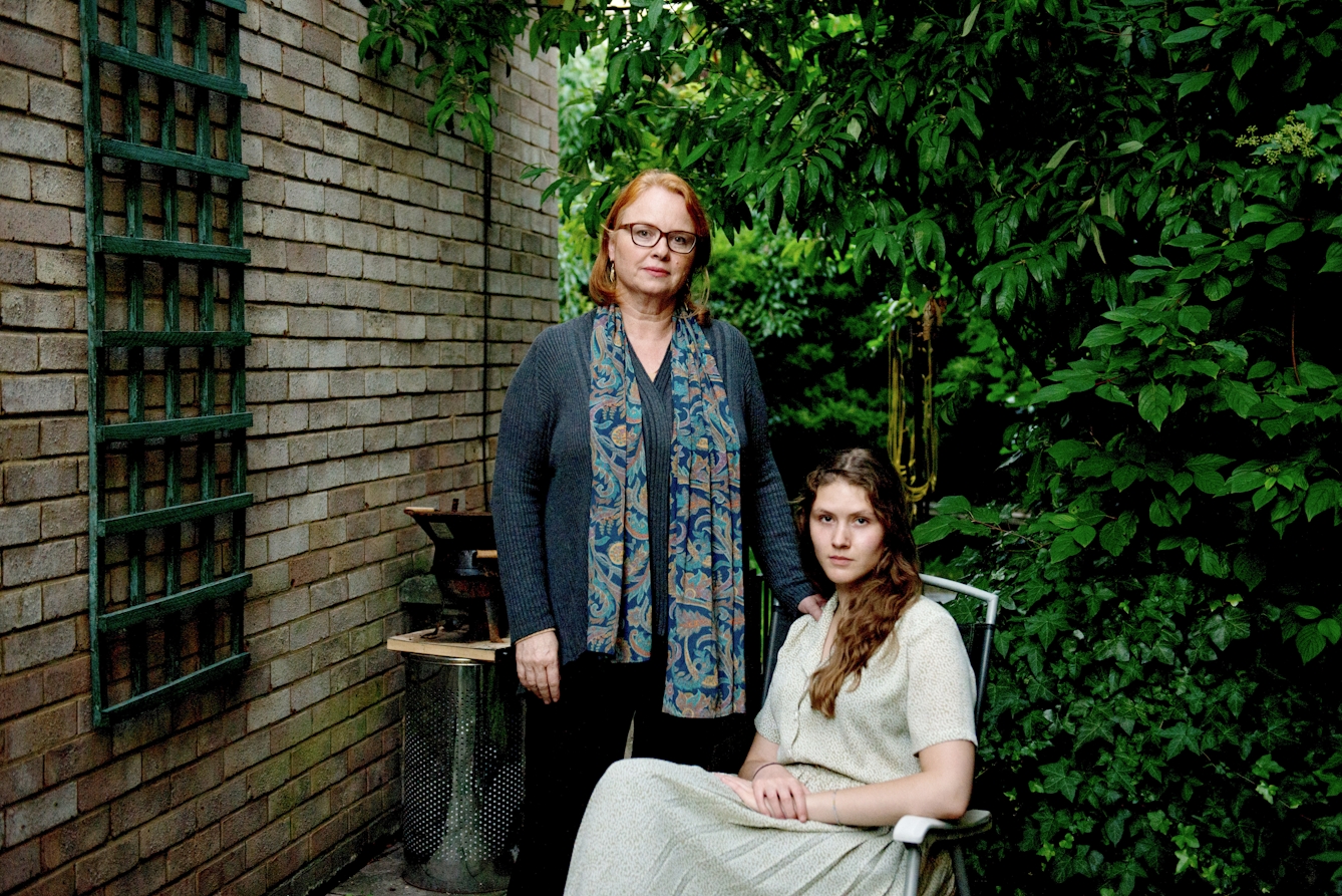 Photograph of a young woman and a mother figure in a domestic garden. The young woman is seated in a long dress and and the mother figure stands behind her, hand on her shoulder. They are both looking to camera.